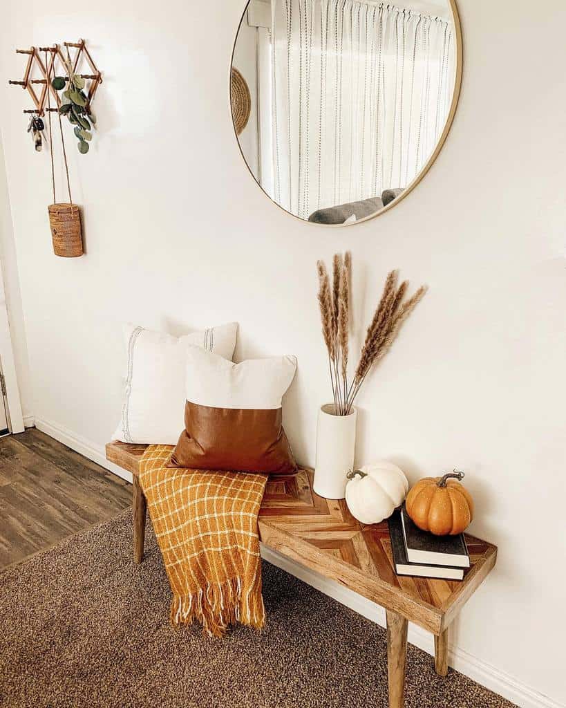 Cozy entryway with a wooden bench, pillows, a plaid throw, pumpkins, books, and a round mirror on a white wall