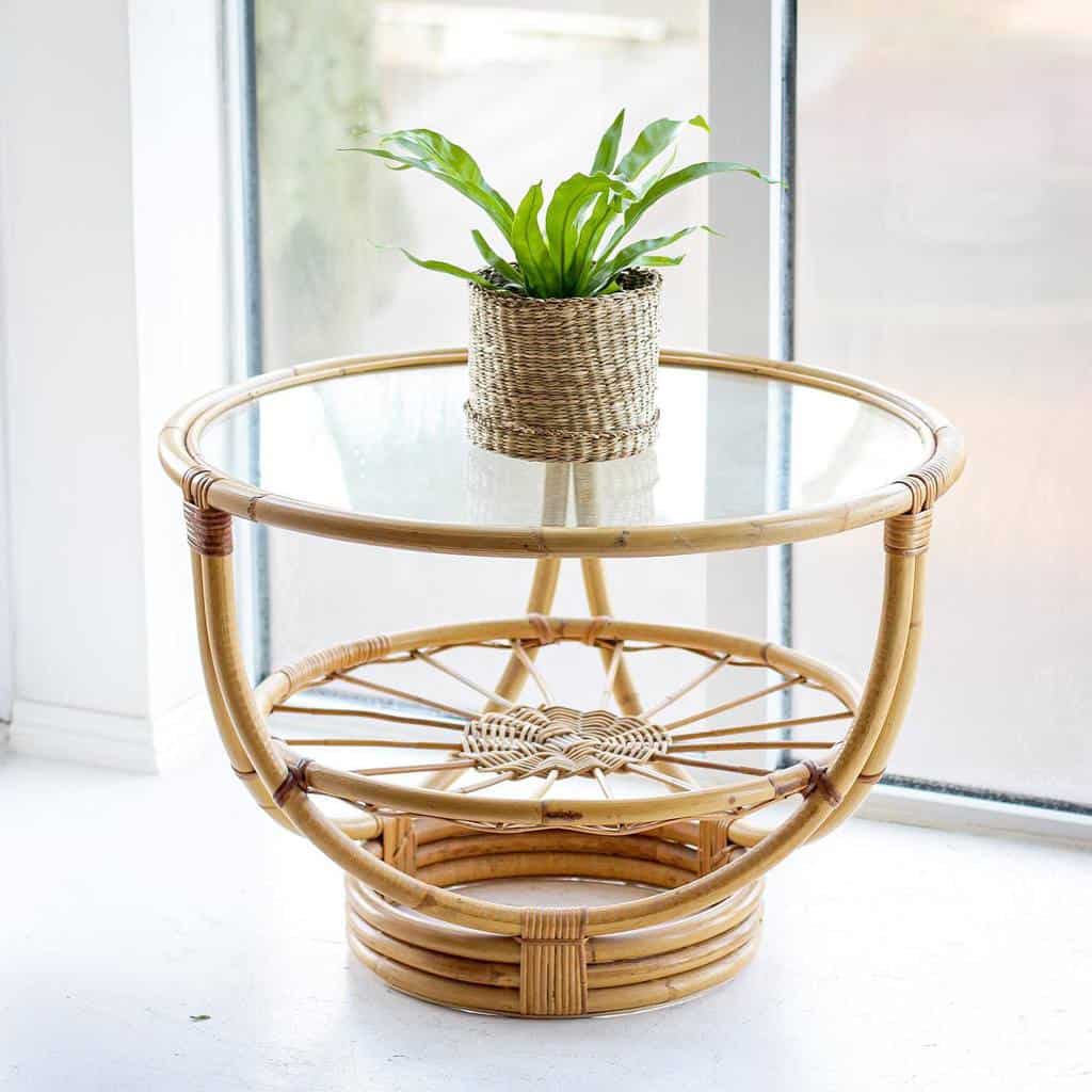 Round glass-topped bamboo coffee table with a woven plant basket, holding a green plant, next to large windows