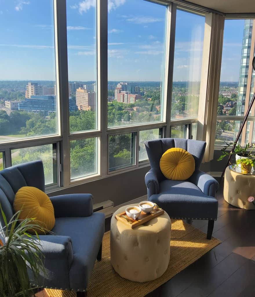 Cozy seating area with two blue chairs, yellow pillows, and a round ottoman with coffee, overlooking a cityscape from large windows