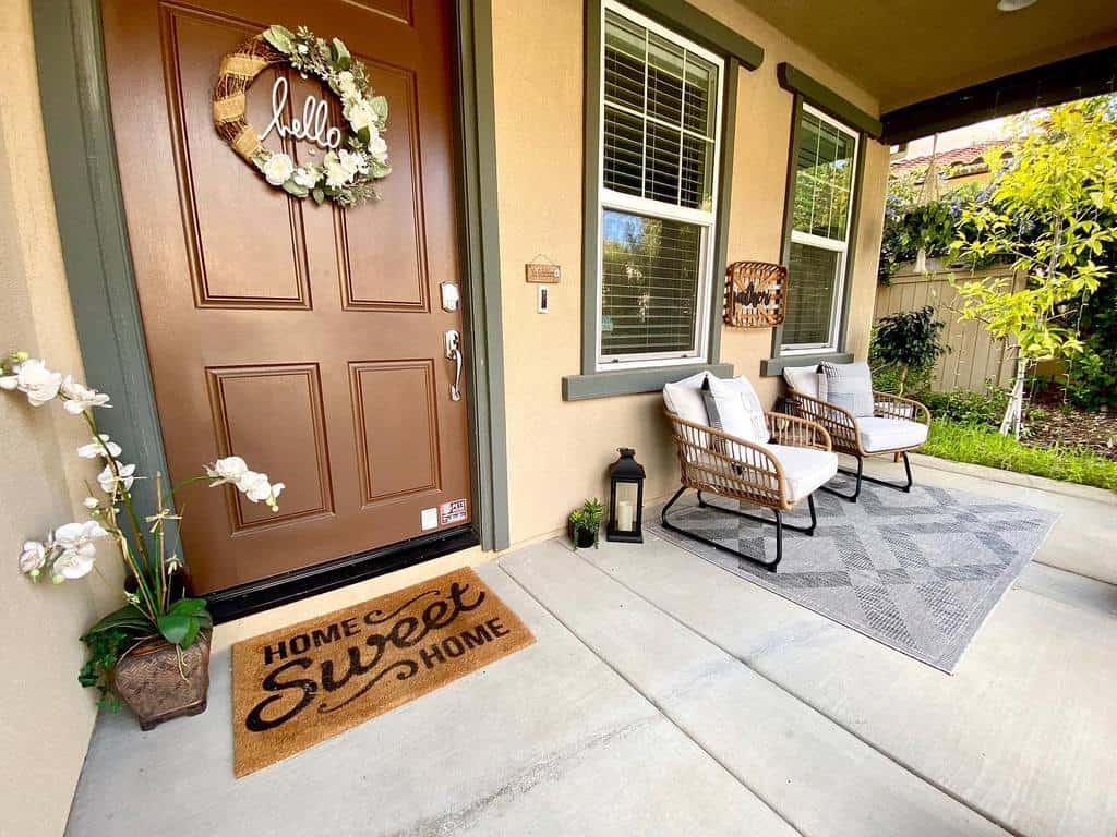 Cozy front porch with two chairs on a rug, a "Home Sweet Home" doormat, and a door with a "hello" wreath, surrounded by plants and lantern decoration