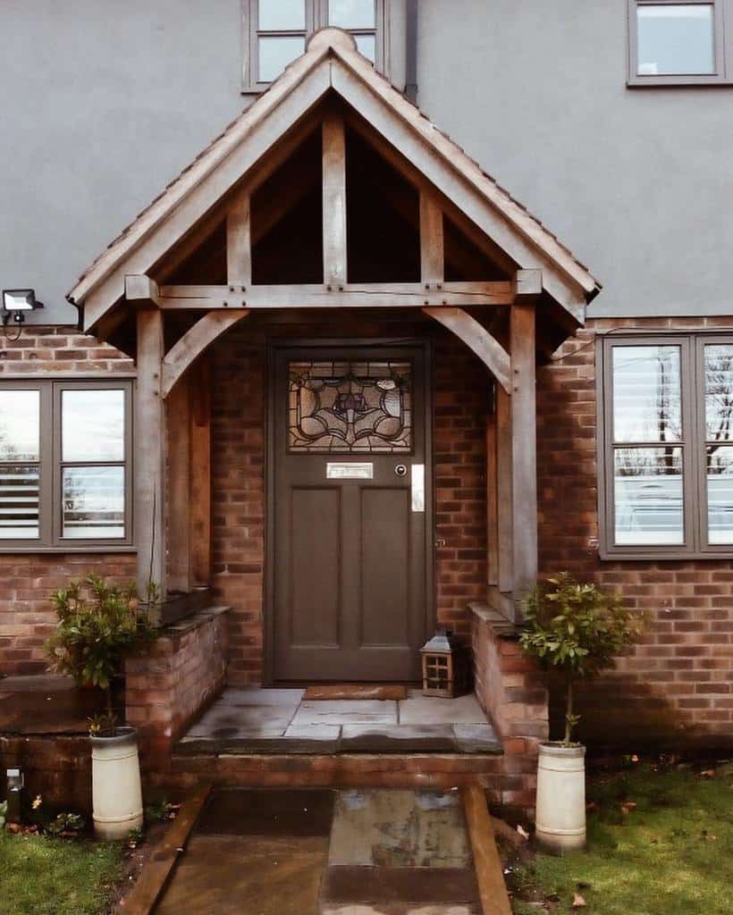 Charming house entrance with a wooden porch, decorative front door, and brick exterior, flanked by potted plants on a stone path