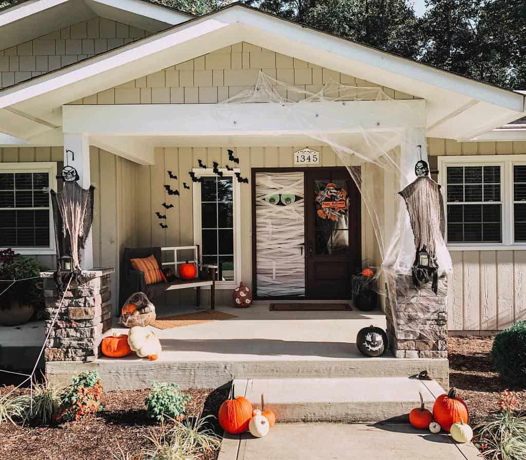 House porch decorated for Halloween with pumpkins, bats, cobwebs, and spooky figures; Mummy-themed door with green eyes