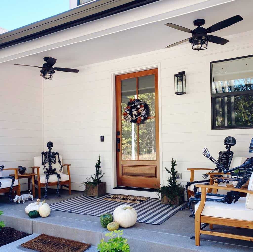 A porch decorated for Halloween with skeletons on chairs, pumpkins on the steps, and a wreath on the wooden door