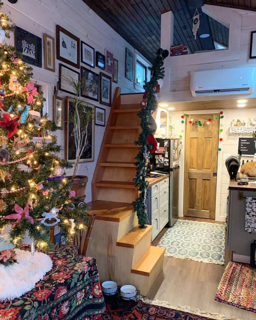 Cozy interior with a decorated Christmas tree, wooden stairs, and art-filled walls. A small kitchen in the background, festive atmosphere.