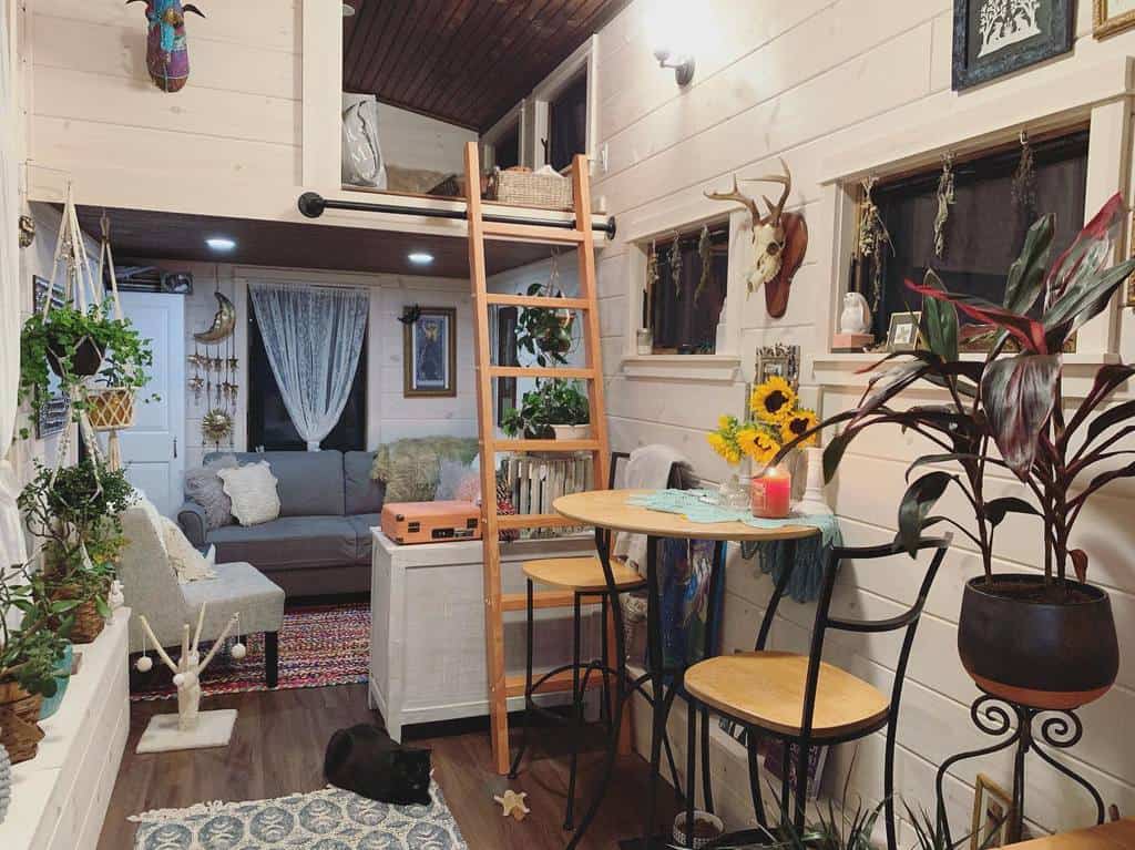 Cozy tiny house interior with plants, a loft ladder, round table with sunflowers, and a sofa; warm lighting and eclectic decor.