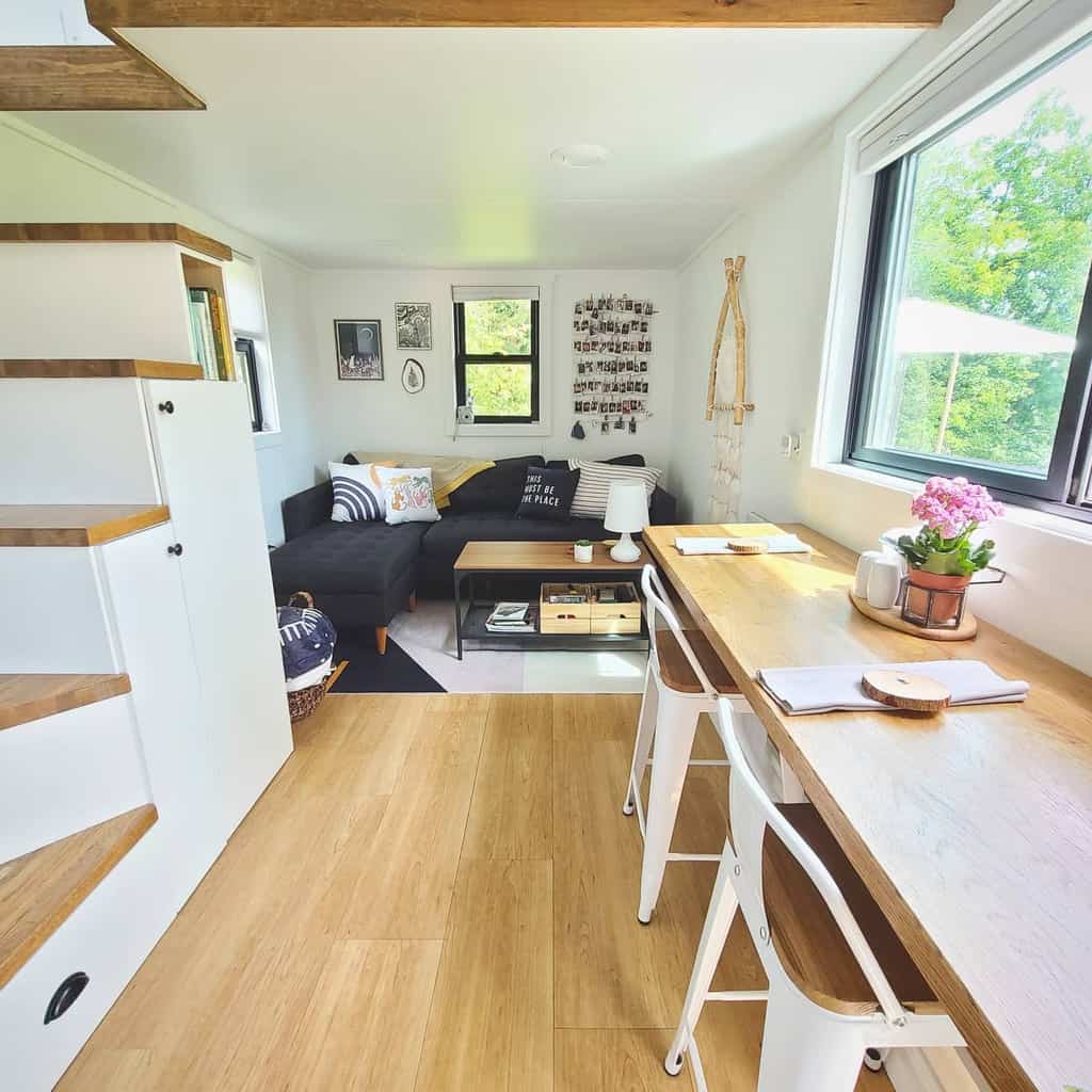 Modern tiny home interior with wooden floors, a small black sofa, wall art, plants, and a wooden table by large windows; bright and cozy.