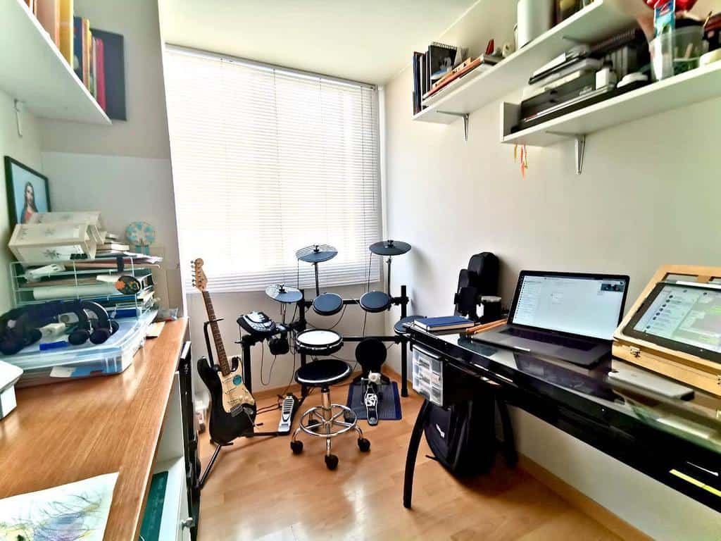 Compact music workspace with an electronic drum set, electric guitar, modern desk setup, floating shelves, and natural light from a large window