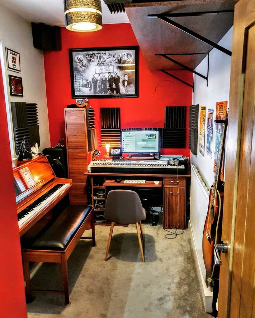 Compact music studio with a piano, keyboard workstation, wall-mounted guitars, acoustic panels, and a bold red accent wall