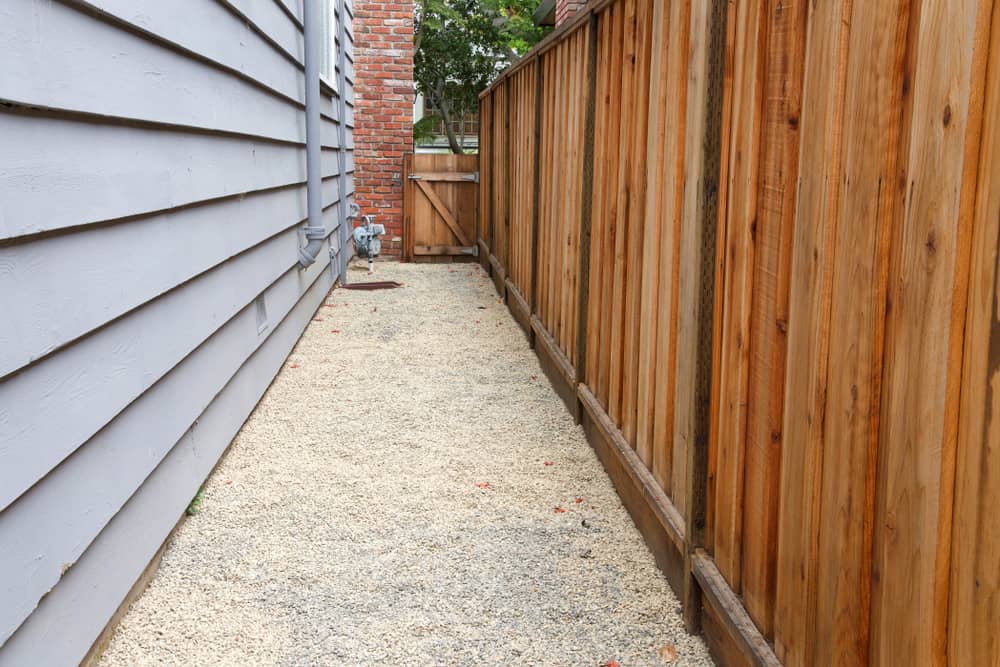 Narrow gravel pathway between a gray wooden house wall and a wooden fence, leading to a wooden gate at the end