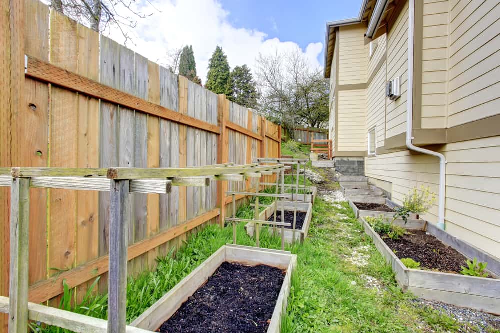 Wooden raised garden beds alongside a house and fence on a grassy path, ready for planting 