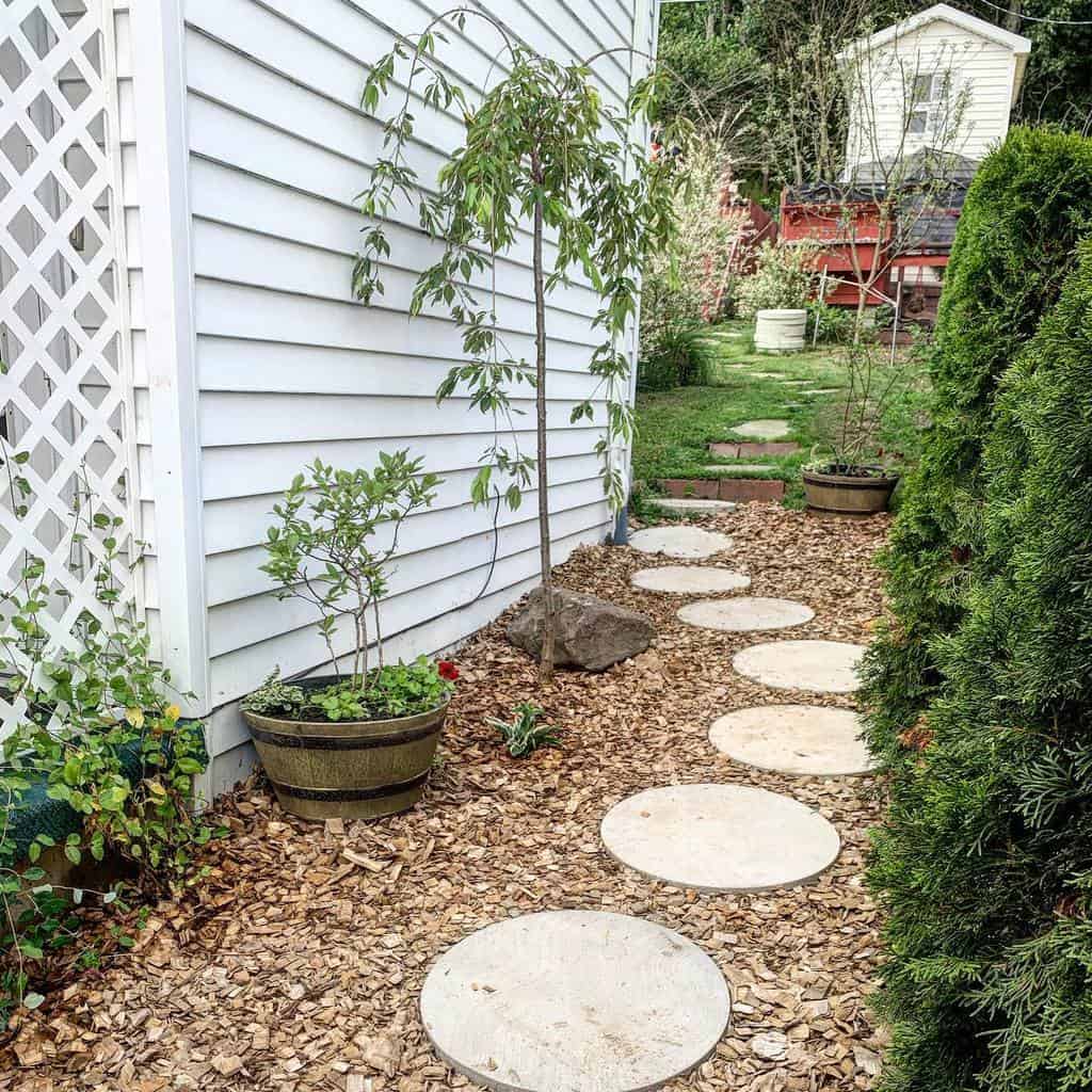 A garden path with circular stepping stones, bordered by plants and a white house wall, leads to a yard with greenery