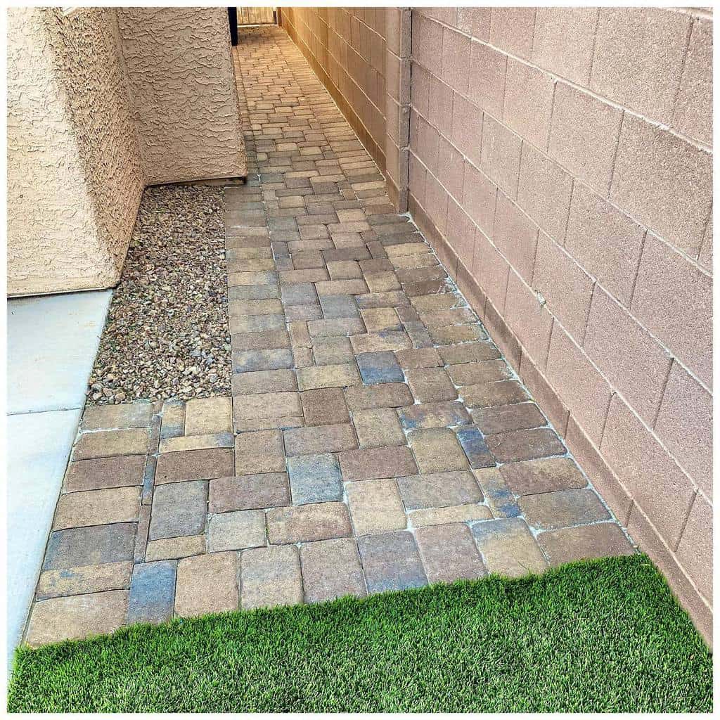 A narrow walkway with stone pavers runs between two walls, bordered by a grassy area and gravel section