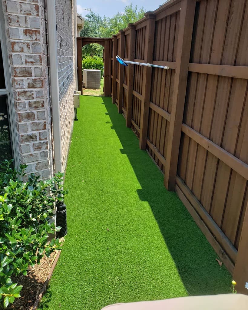 Narrow side yard with artificial green grass, bordered by a brick house and a wooden fence, leading to a gate