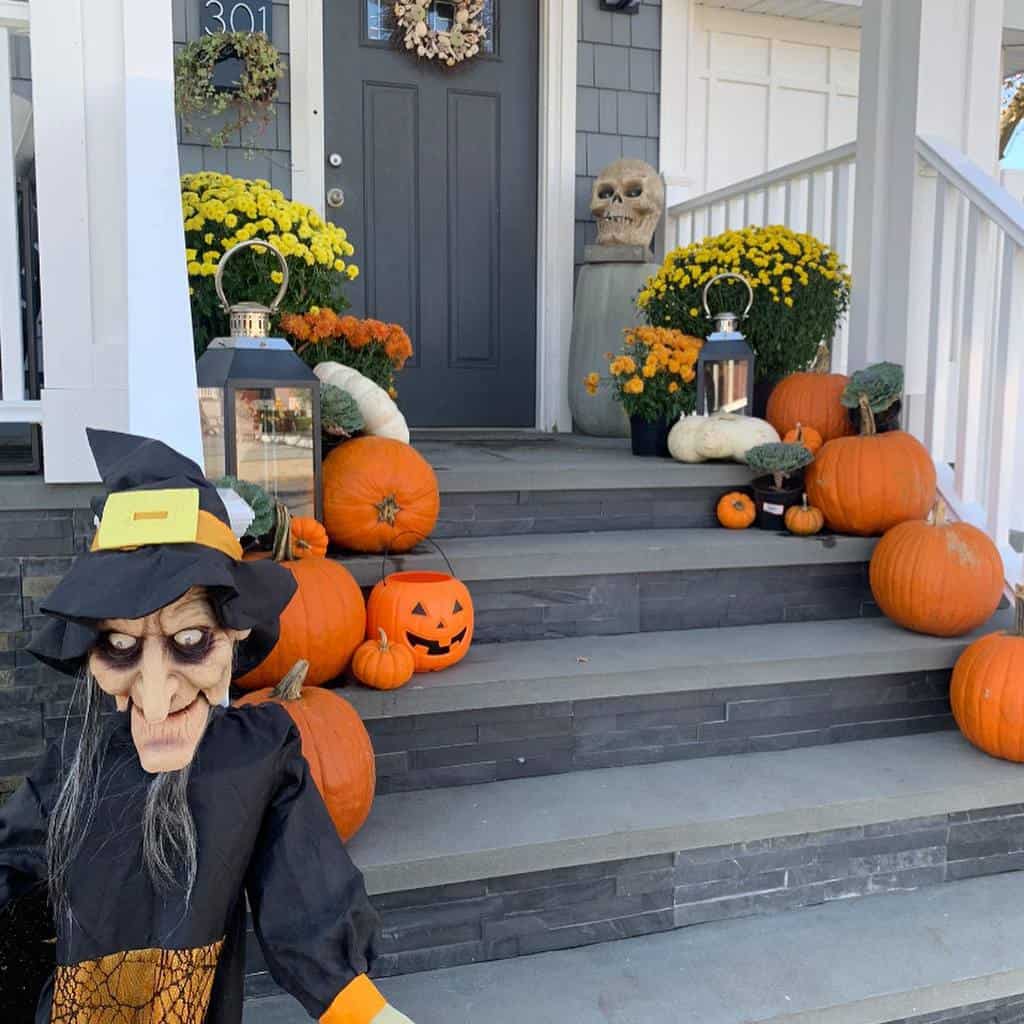 A porch decorated for Halloween with pumpkins, a witch figure, lanterns, autumn flowers, and a skull by the steps