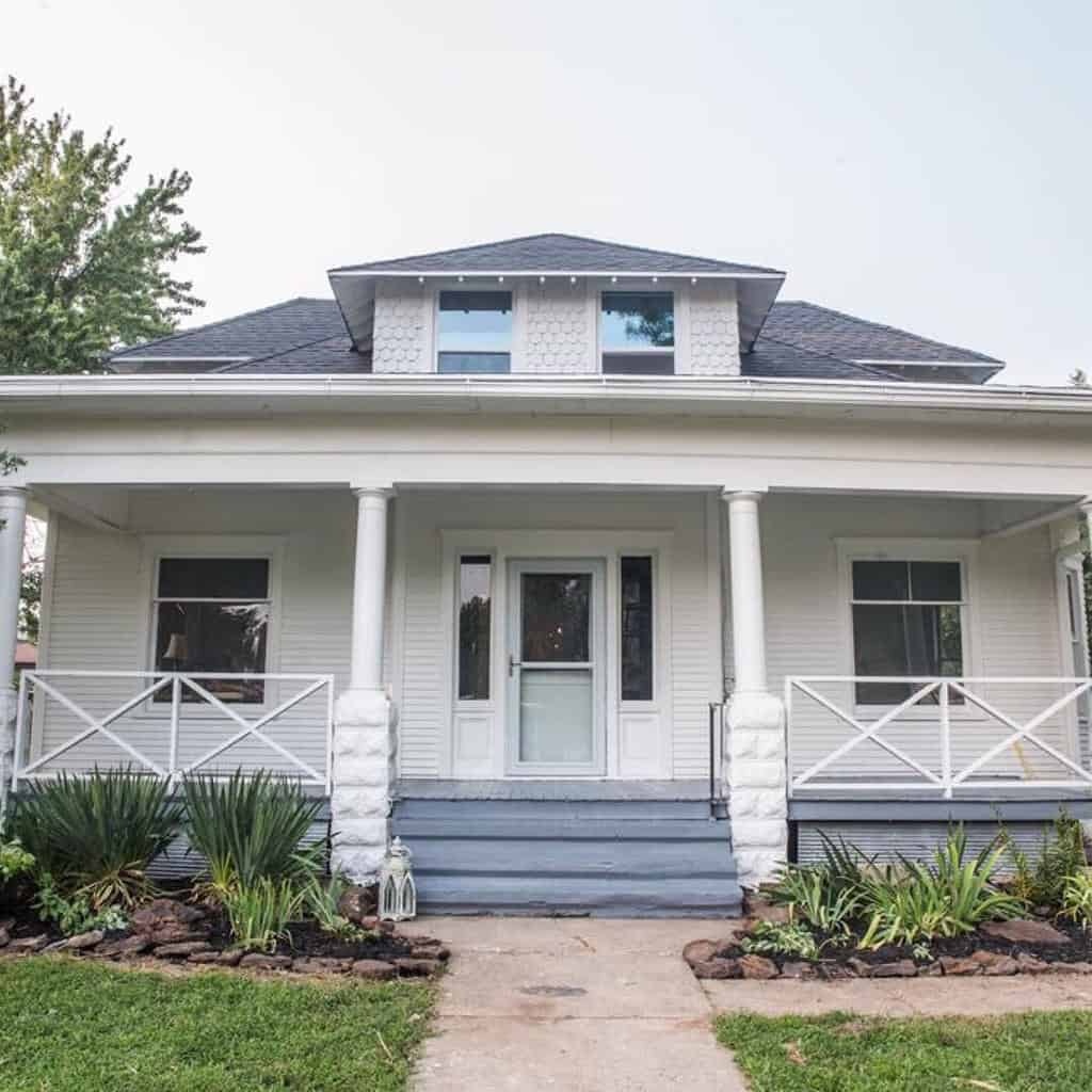 A white bungalow house with a front porch, symmetrical windows, and a path leading to the entrance, surrounded by greenery and plants