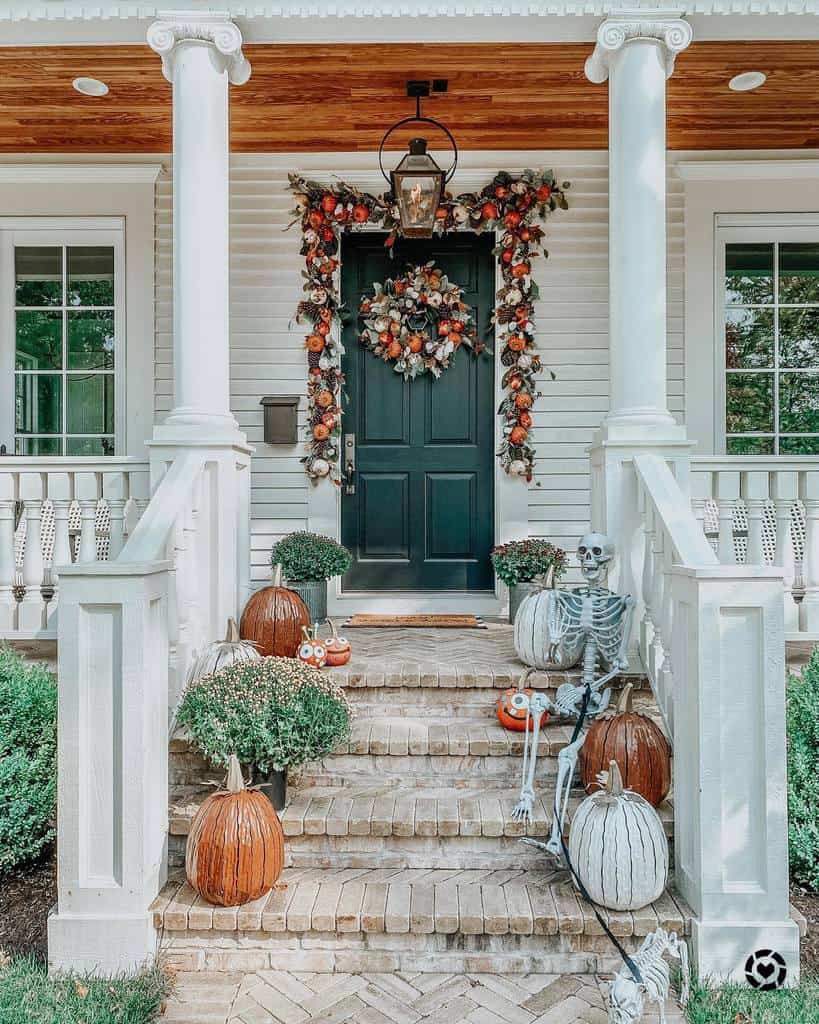 Front porch with autumn decorations: a black door with a fall wreath, pumpkins, a skeleton sitting on steps, and potted plants