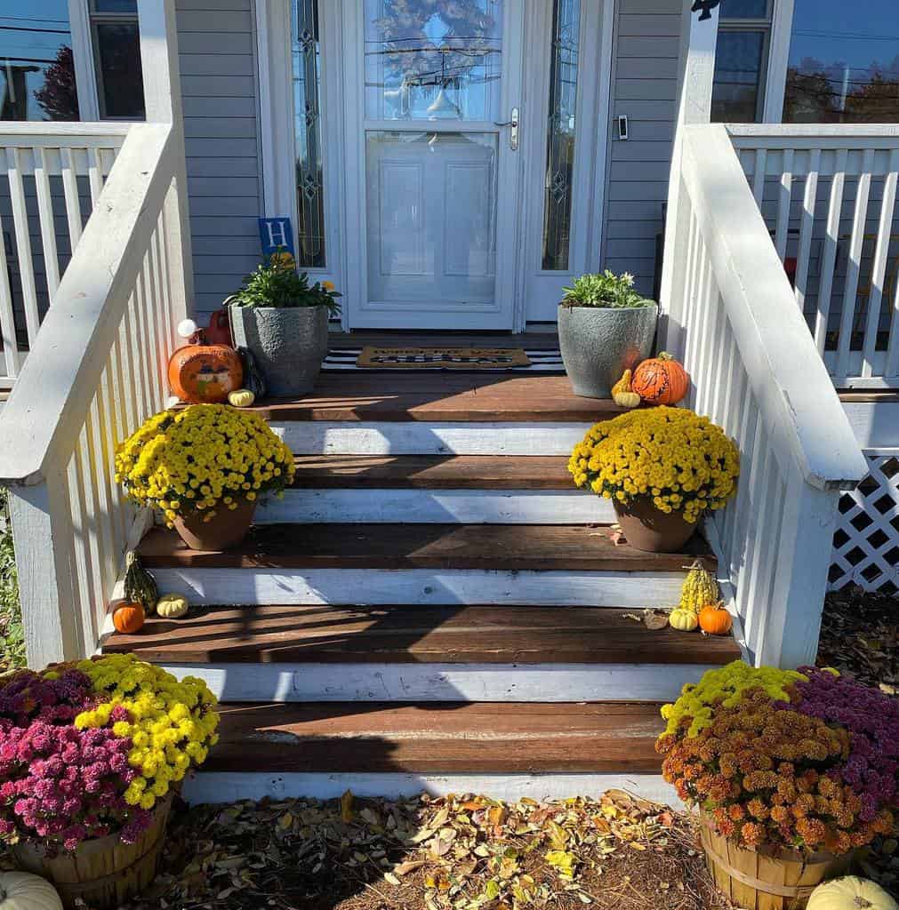 House entrance with white railings, potted yellow flowers, pumpkins, and gourds on the steps under a sunny sky