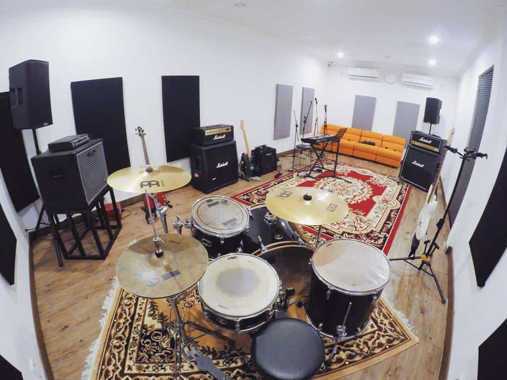 Spacious music studio with a drum set, guitars, amplifiers, a keyboard, acoustic panels, and a cozy orange sofa on a patterned rug