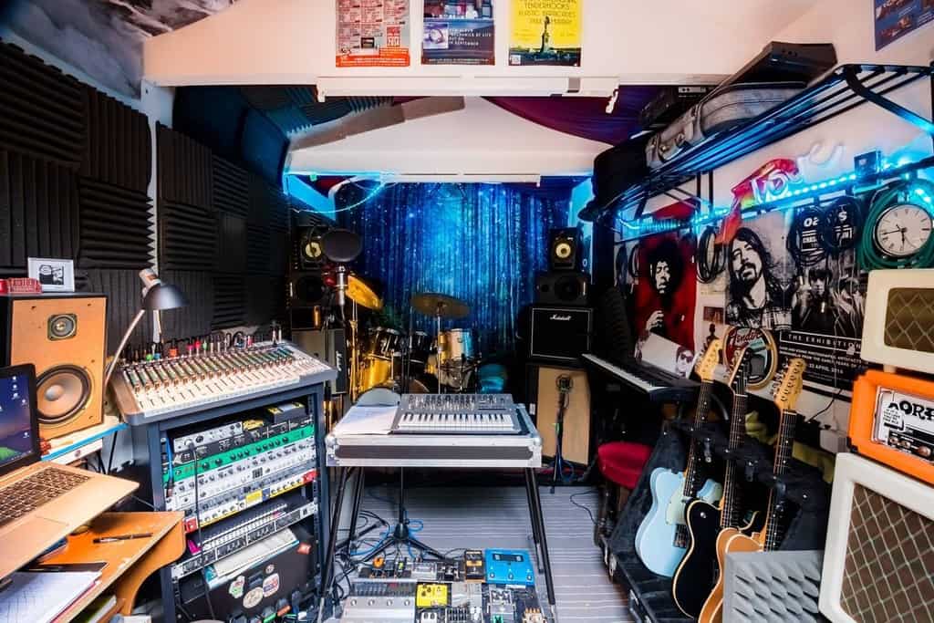 Eclectic home music studio with guitars, a drum set, synthesizers, amplifiers, a mixing console, acoustic panels, and vibrant LED lighting