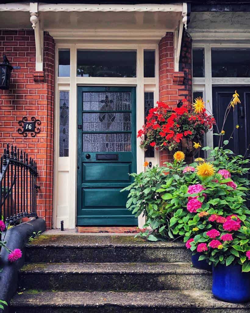 Brick house entrance with a dark green door, flower pots with colorful blooms on either side, and steps leading up to the door