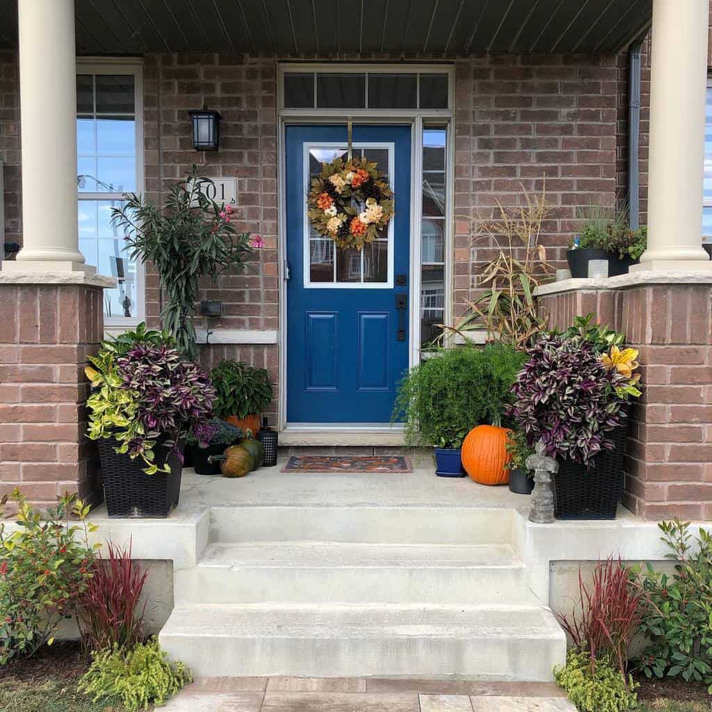 A blue front door with a floral wreath, surrounded by potted plants and pumpkins on a brick porch with steps