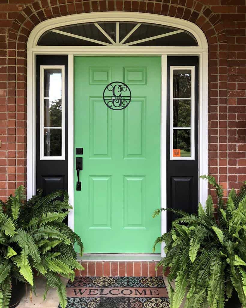 A vibrant green door with black trim, surrounded by red brick, flanked by windows, and adorned with potted ferns and a welcome mat