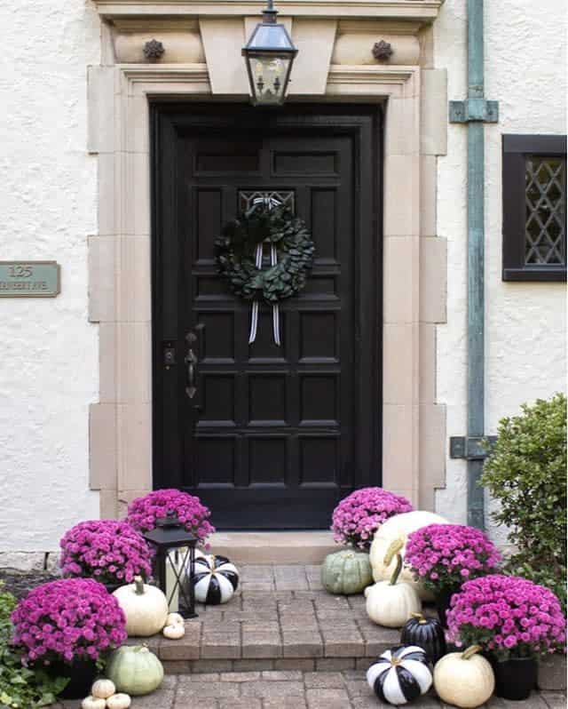 A black door with a wreath, surrounded by pink flowers and black-and-white pumpkins on the steps, lantern above the door