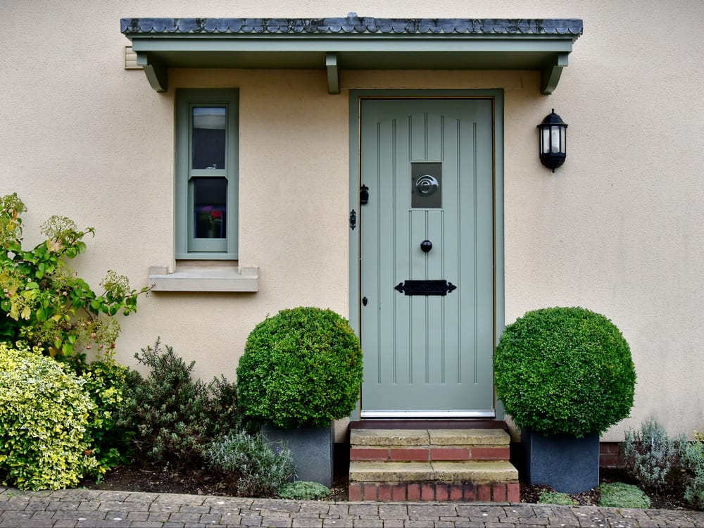 A light green door with a small window and a black lantern on a cream house, two round bushes in pots flank the entrance