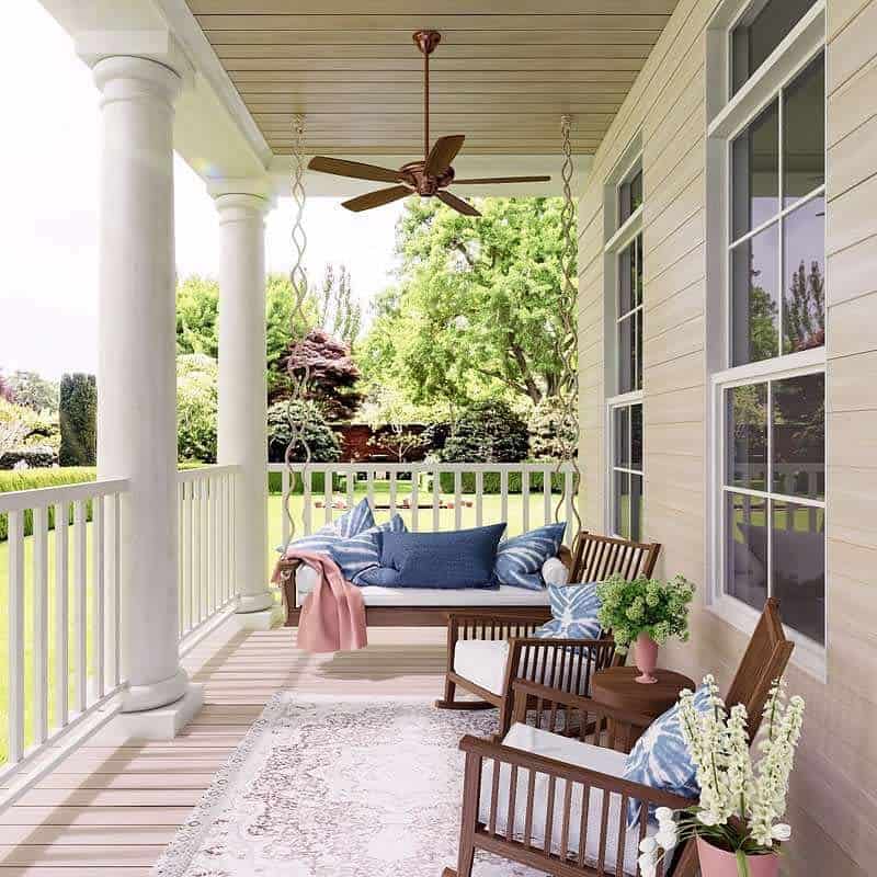 A cozy front porch with a swing, chairs, cushions, plants, and a fan overlooks a green garden through white railings