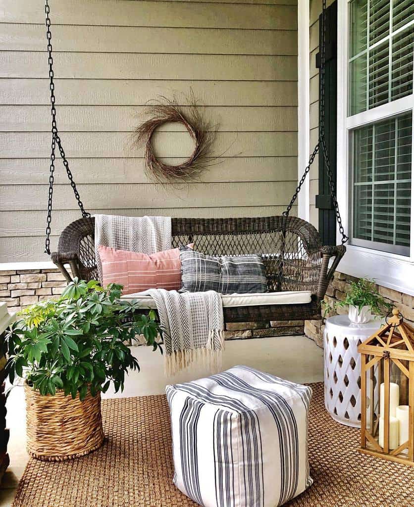 Cozy front porch with a wicker swing, patterned cushions, plants, lanterns, and a wall wreath