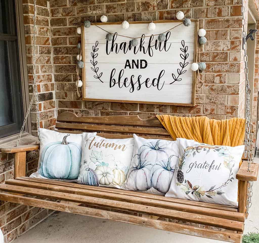 Wooden swing with fall-themed pillows and "Thankful and Blessed" sign on a brick wall, string lights add a cozy touch