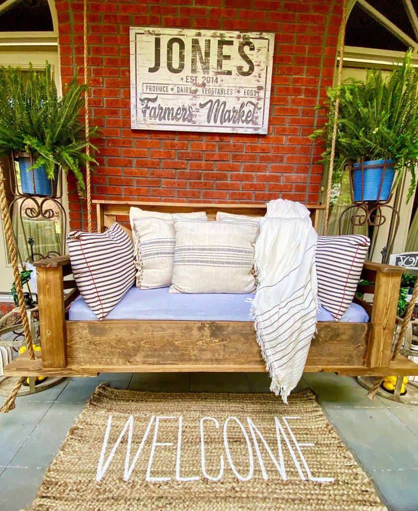 Cozy porch bench with striped pillows and a throw blanket, under a "Farmers Market" sign, surrounded by green ferns, "Welcome" mat in front