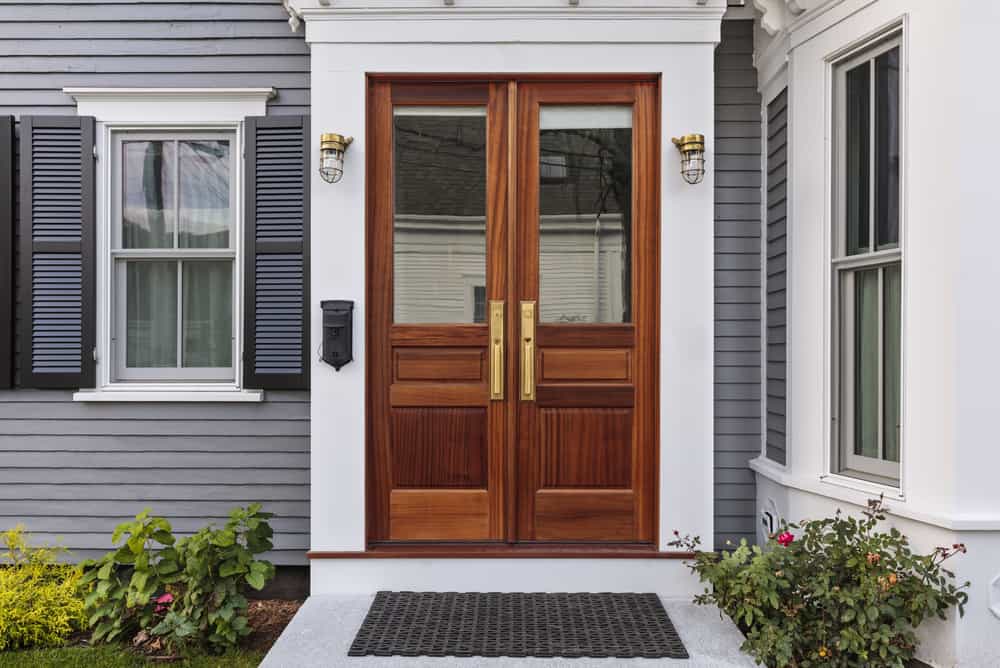 Elegant wooden double front doors with glass panels, set in a gray house with white trim, flanked by shutters and small garden plants