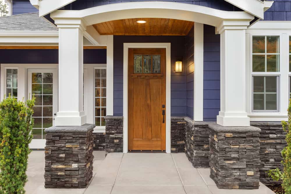 Front porch of a modern house with a wooden door, stone-accented columns, and blue siding