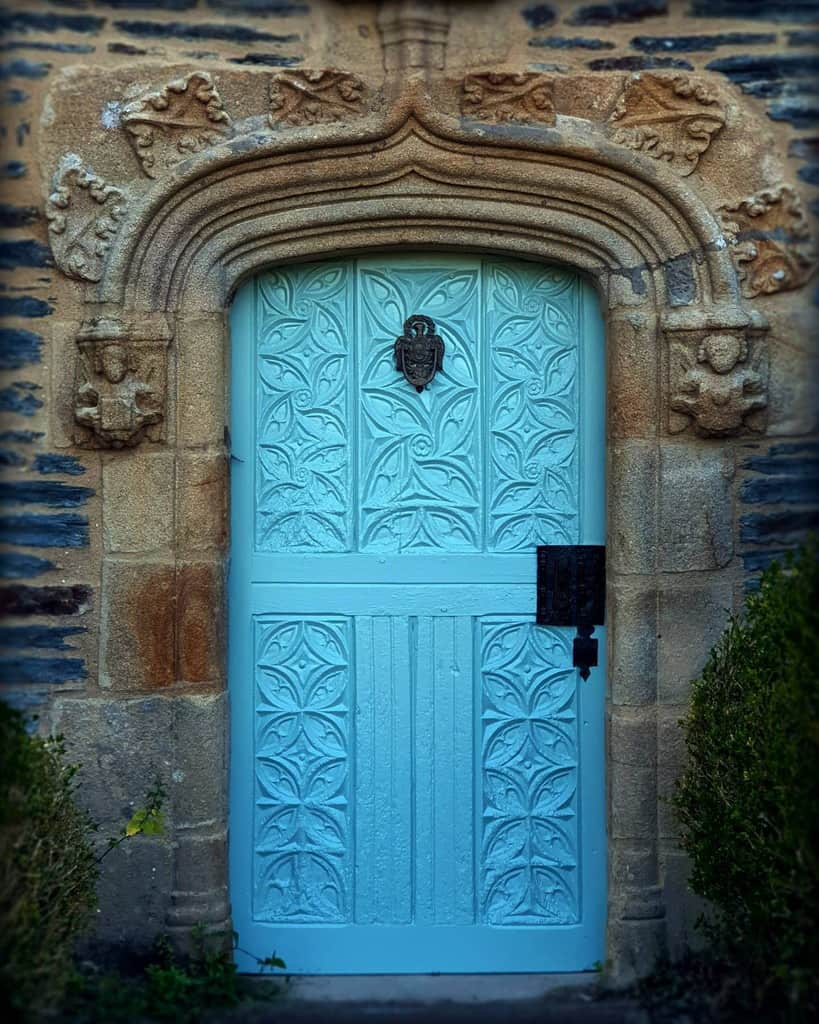 Ornate blue door with intricate carvings, set in a stone archway, featuring a decorative knocker and surrounded by greenery