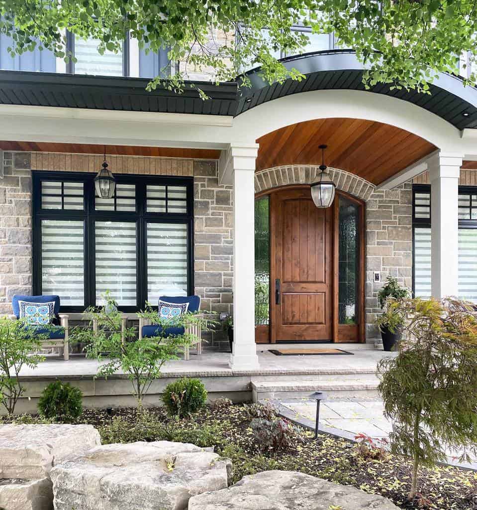 Modern house entrance with a wooden door, stone facade, two chairs with blue cushions, and surrounding greenery