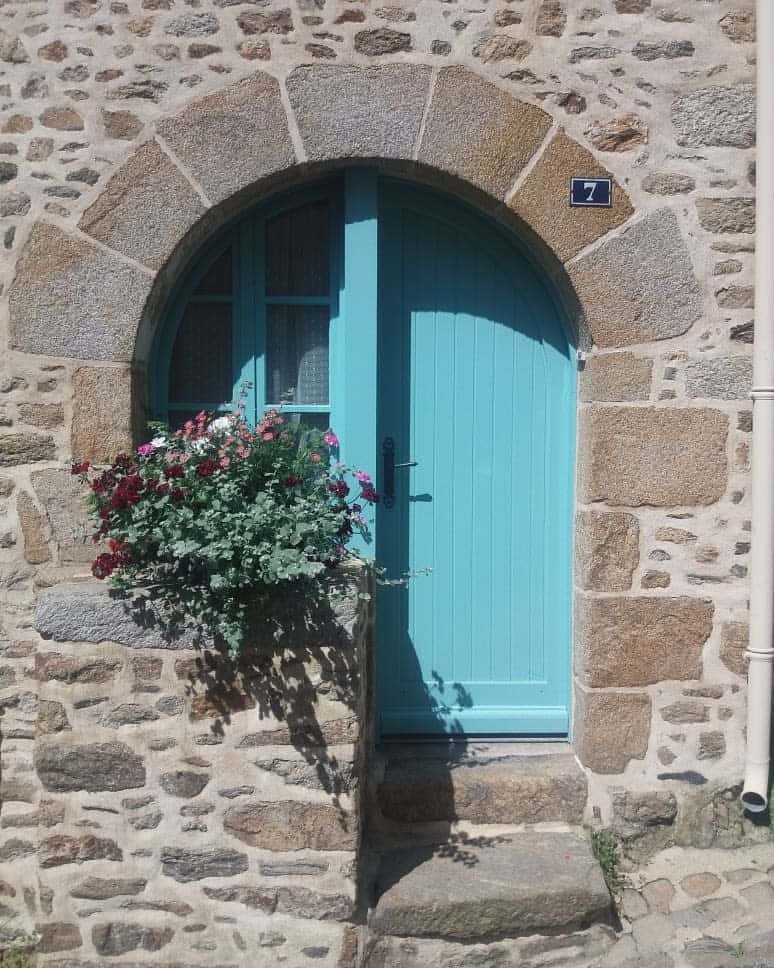 Stone wall with an arched, bright blue door and a small flower box with red and white flowers below house number 7