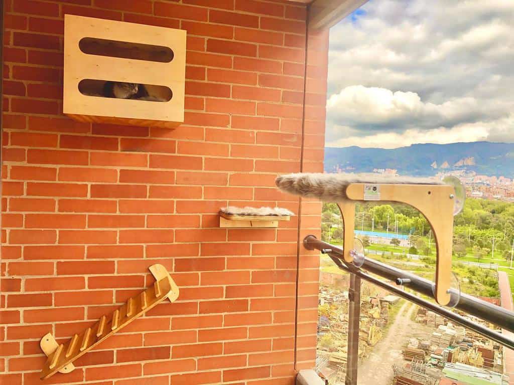 A covered balcony with a wall-mounted cat box, a small platform, and a ladder. A view of the city and mountains is visible outside