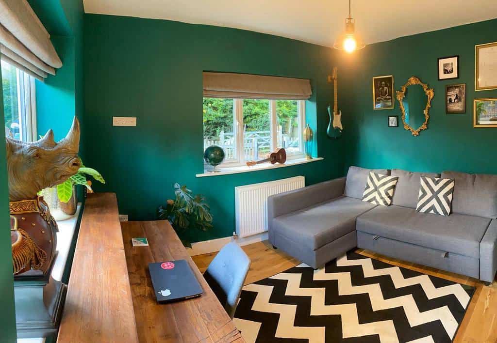 Stylish music room with deep green walls, a wall-mounted guitar, a cozy gray sofa, a wooden desk, and bold black-and-white decor accents