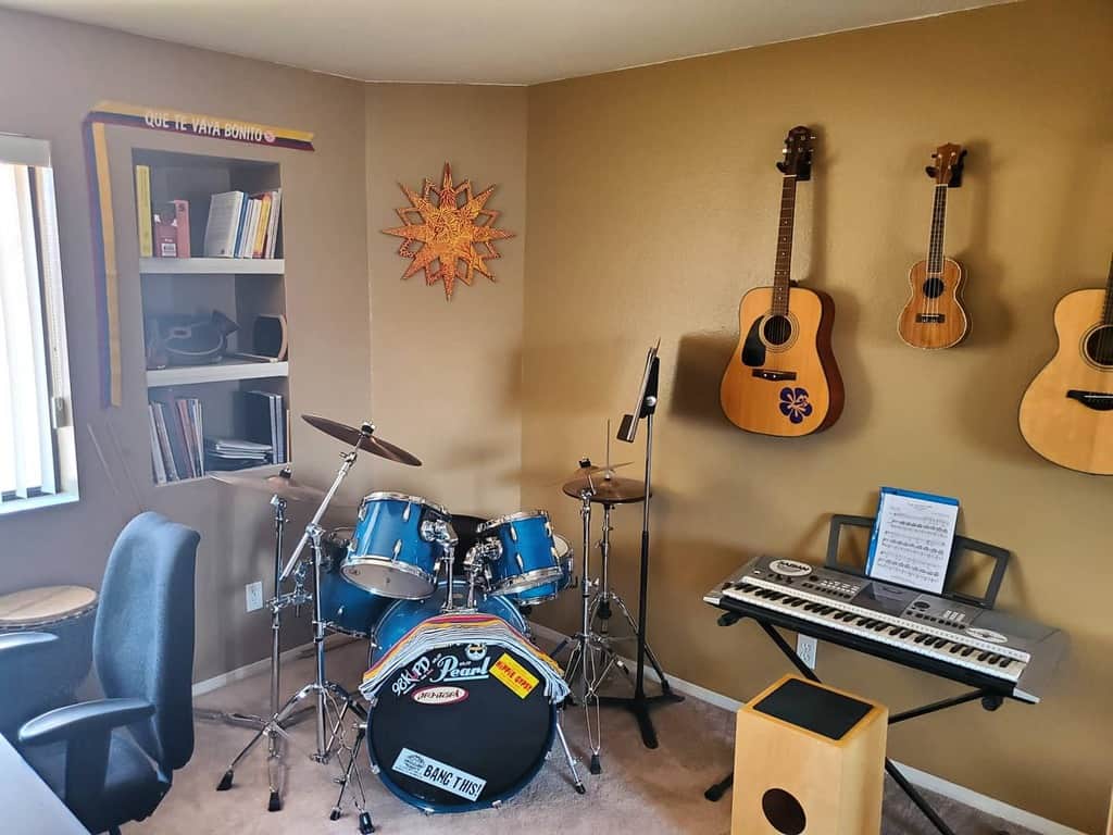 Cozy home music room with warm-toned walls, wall-mounted guitars, a drum set, a keyboard, a cajón, and a built-in bookshelf