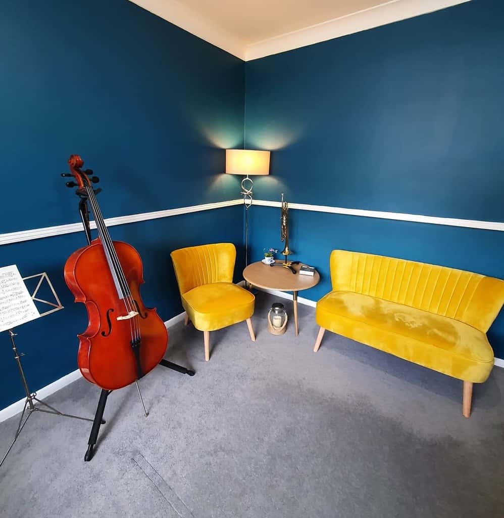 Elegant music room with deep blue walls, mustard yellow seating, a cello, a trumpet, soft lighting, and a cozy minimalist design