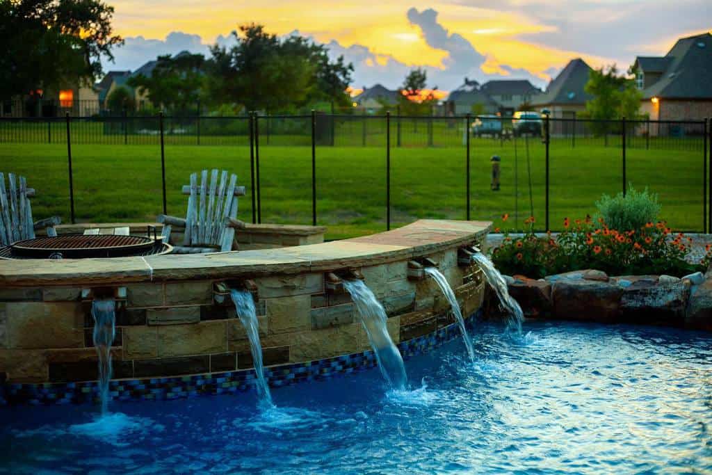 Backyard pool with cascading waterfalls, surrounded by chairs and flowers, with a scenic sunset and fence with houses visible in the background