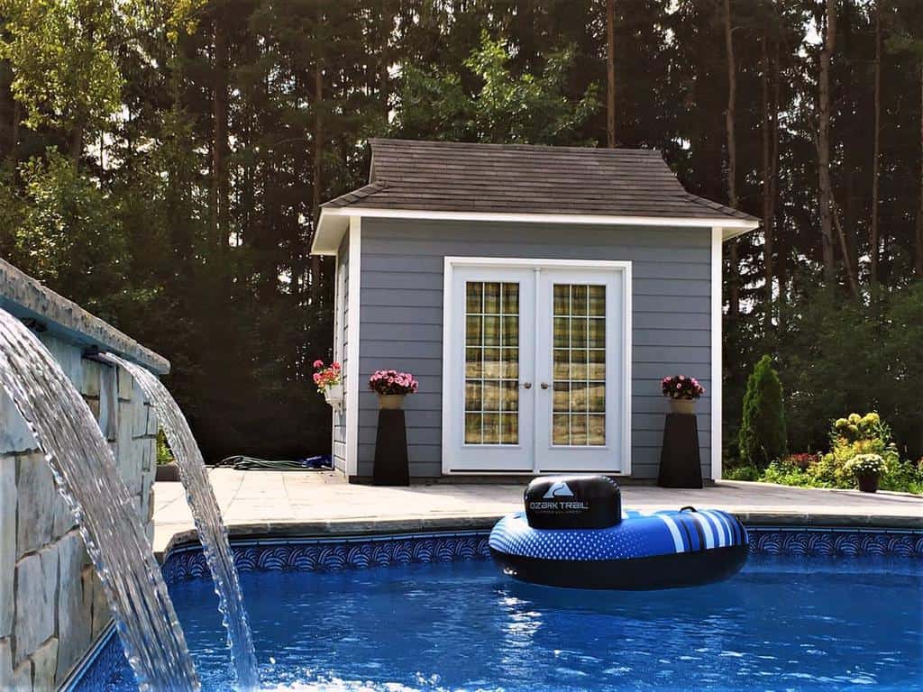 Poolside view with blue and white inflatable ring, waterfall feature, and a small grey pool house in the background