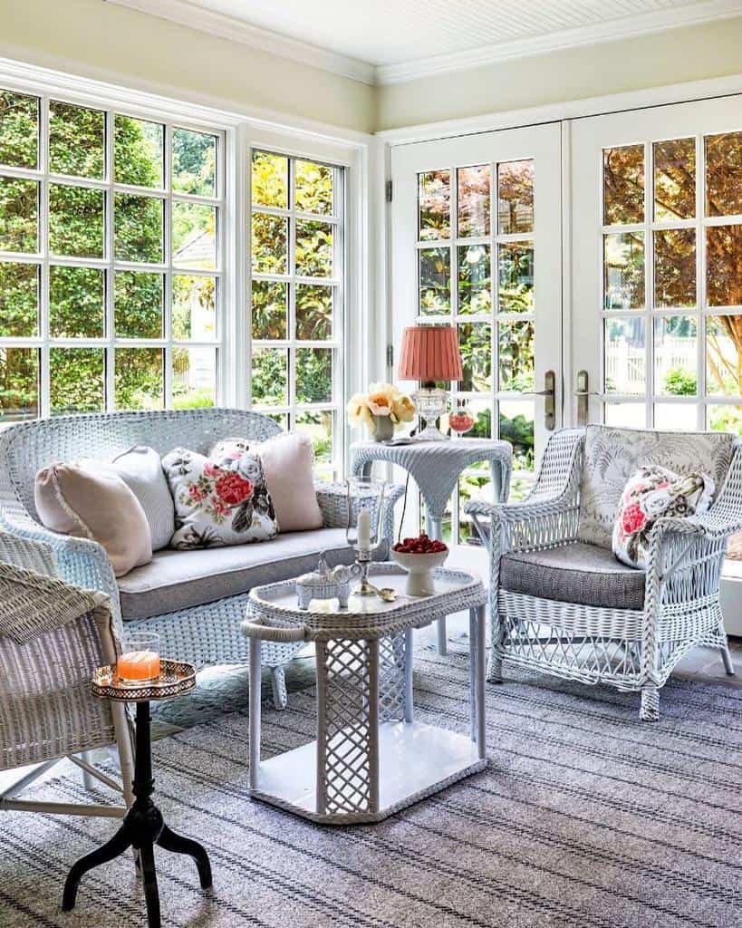 Sunlit room with white wicker furniture, floral cushions, a small table with teapot, and large windows overlooking a garden