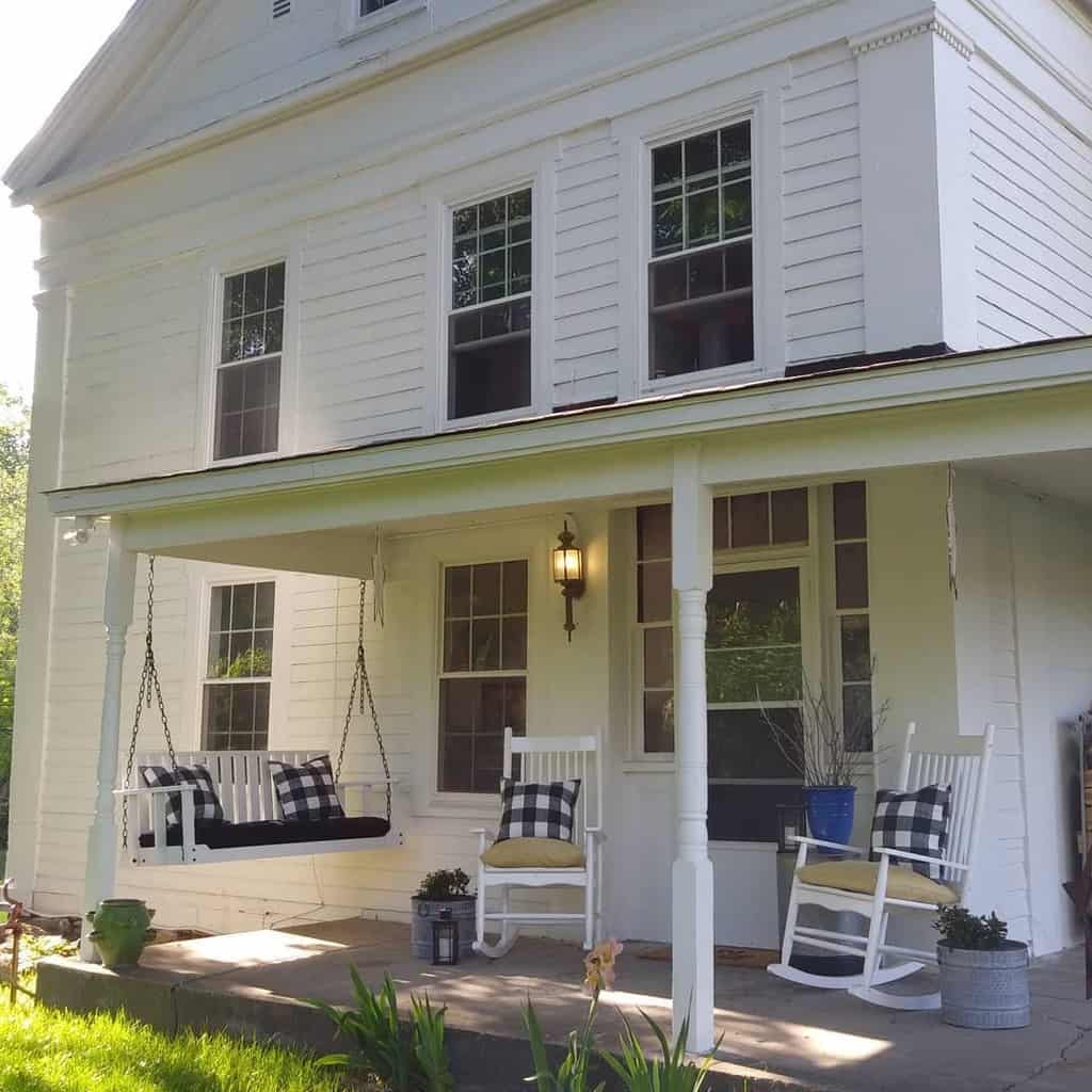 A white house with a porch swing and rocking chairs, featuring black and white plaid cushions, and potted plants by the entrance