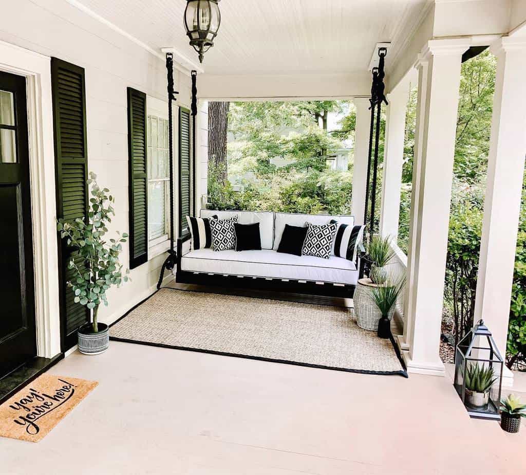 A cozy porch with a white swing, black-and-white cushions, potted plants, and a "Yay! You're Here!" doormat, creating a pleasant and inviting atmosphere