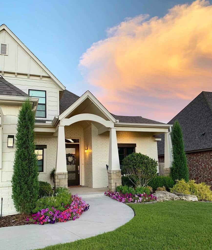 A modern house with a gable roof, surrounded by vibrant flowers and green lawn under a colorful sunset sky