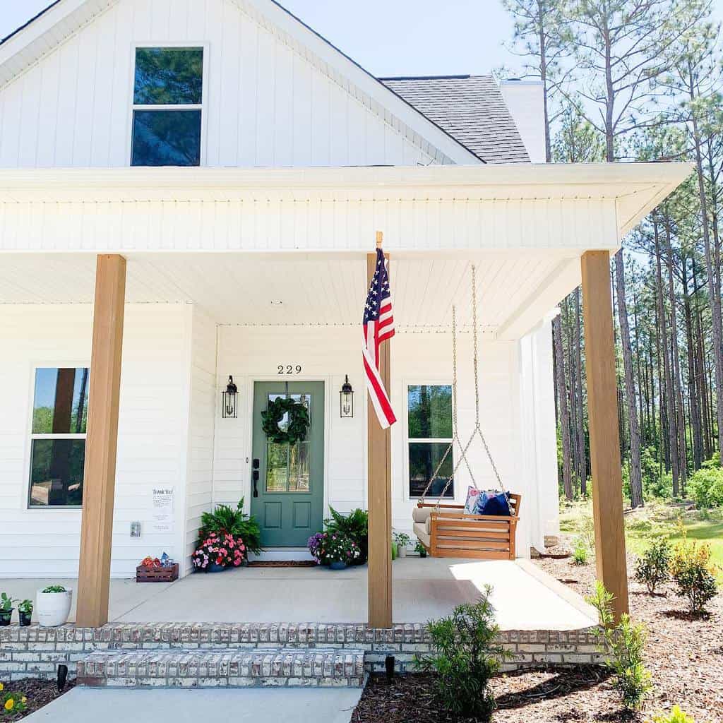 White house with green door, American flag, porch swing, lanterns, and plants. Brick steps lead to the entrance