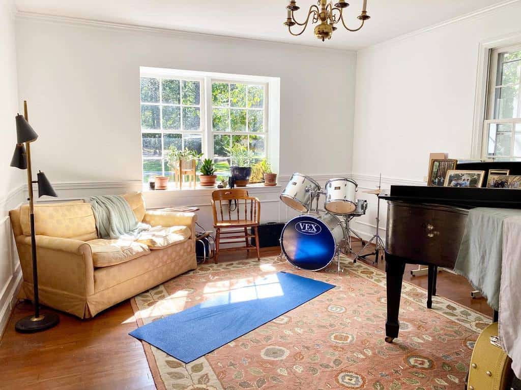 A cozy music room with a sofa, drum set, piano, and a blue yoga mat on the floor, sunlight streaming through a large window with plants