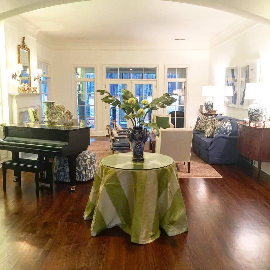 Elegant living room with a grand piano, large round table with floral centerpiece, sofas, and decorative lamps on hardwood flooring